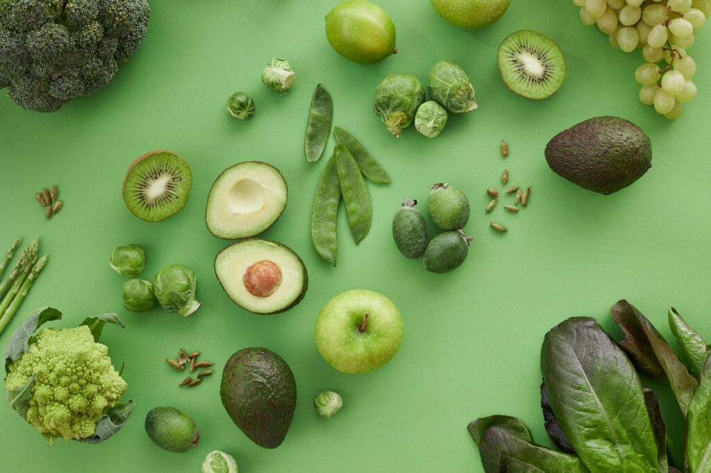 A variety of fresh green vegetables and fruits including avocado, broccoli, kiwi, apples and leafy greens representing a diet that supports gut health