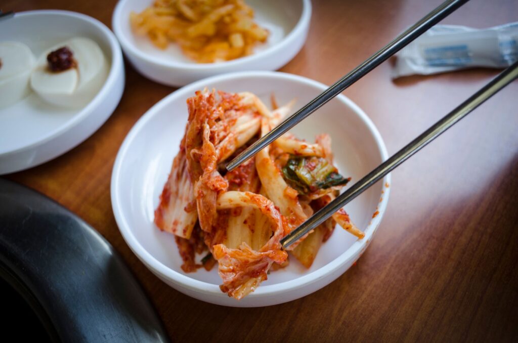 Close-up of traditional Korean kimchi being picked up with chopsticks from a ceramic bowl, representing fermented foods as natural sources of probiotics for gut health