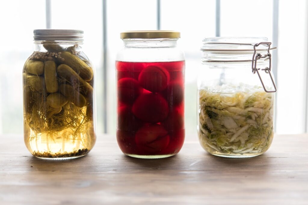 Three glass jars of fermented foods including pickles, pickled beets and sauerkraut on a wooden surface.